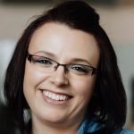 A close-up photo of author, Rebecca Cole. She is smiling at the camera, with white skin, dark red hair and thin framed glasses