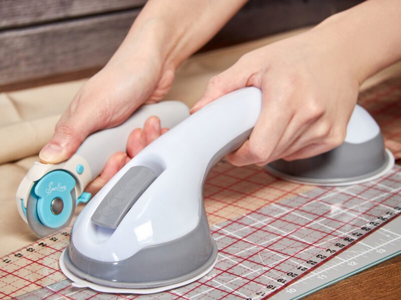 a close up of hands holding a large white and grey suction handle on a grided ruler, cutting with a rotary cutter with their other hand